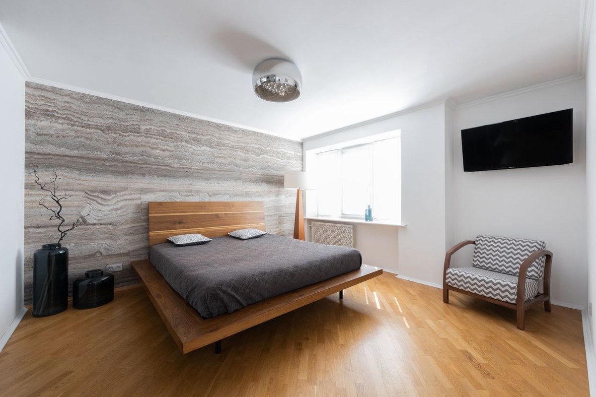 Modern bedroom with a low wooden platform bed, grey quilt, textured accent wall, and chevron armchair