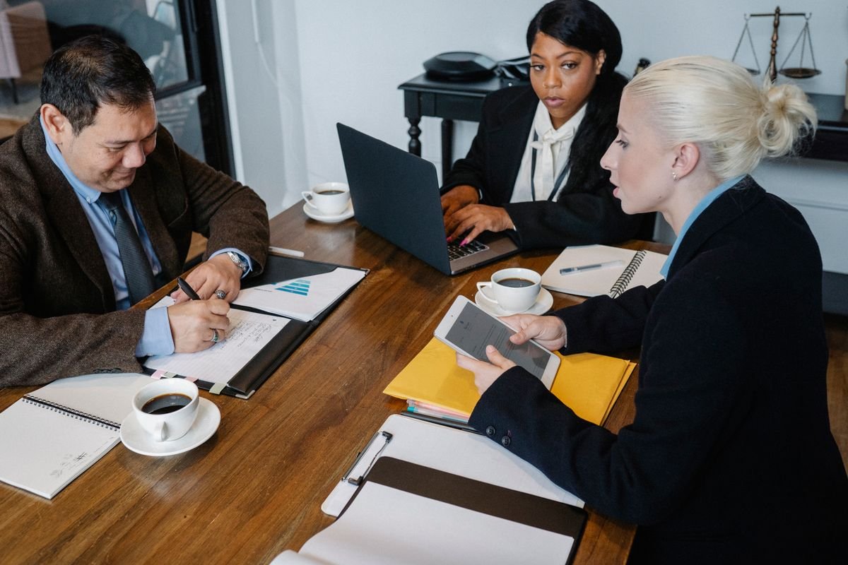 Three colleagues at a wooden meeting table reviewing documents, a laptop and a tablet over coffee
